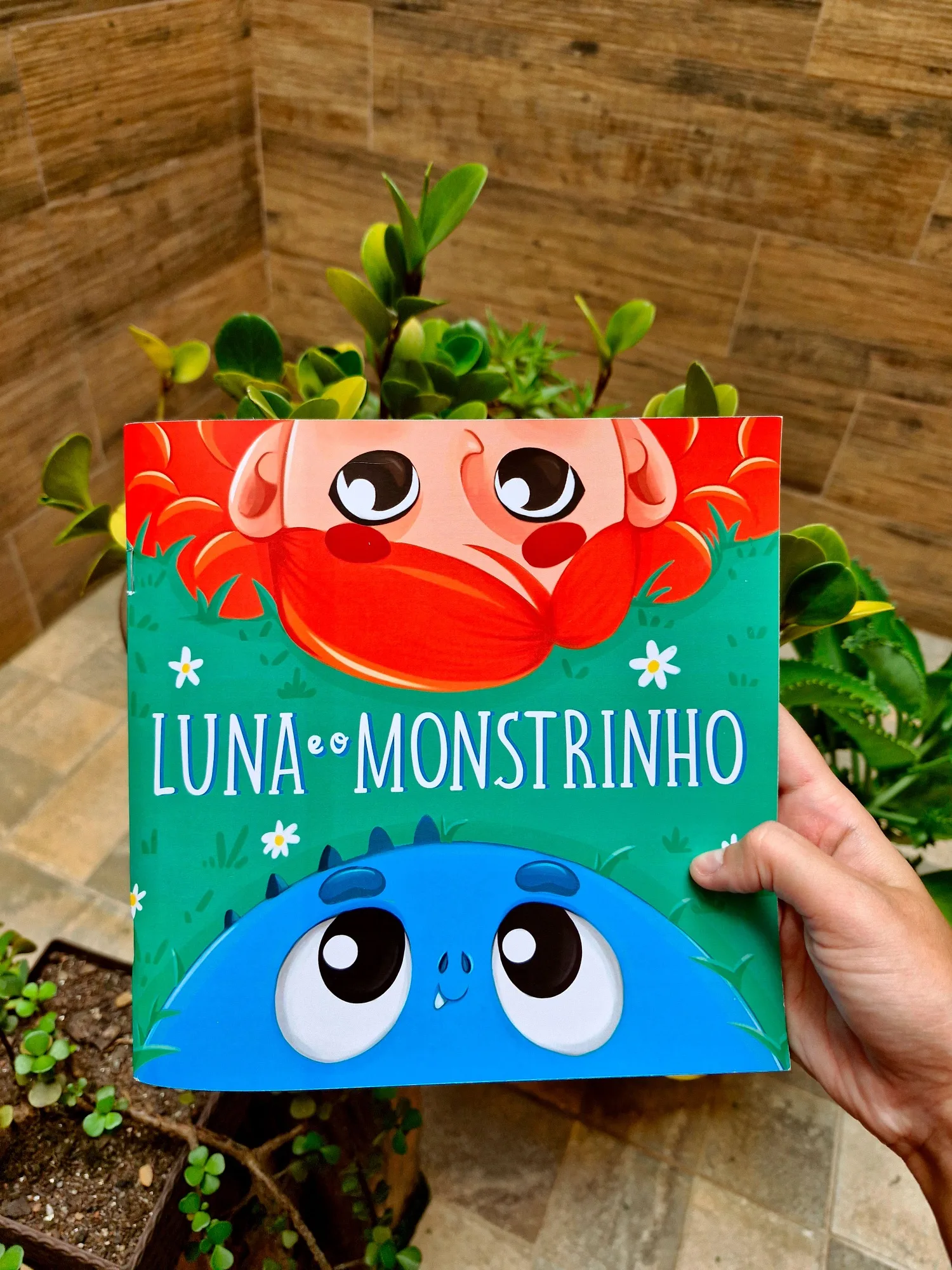 Livro Luna e o Monstrinho impresso sendo segurado na mão, com fundo de plantas